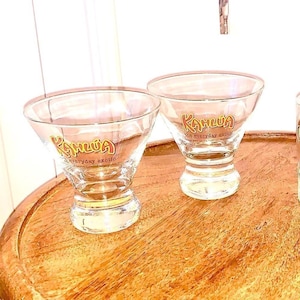 May include: Two clear Kahlúa cocktail glasses with a flared bowl and a short, rounded stem. The Kahlúa logo and "the everyday exotic" are printed in gold on the front of each glass. The glasses are on a wooden surface.