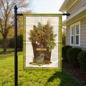 Puede incluir: Una bandera rectangular para exteriores con una ilustración detallada de un cubo rebosante de piñas y vegetación. Un pequeño pájaro está posado debajo. La bandera tiene un borde verde y se muestra en un poste de metal negro.