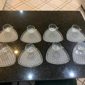 May include: Eight clear glass teacups with matching triangular saucers. The saucers have a textured, dotted pattern. The teacups sit in a circular indentation on the saucers. The set is arranged on a dark countertop.