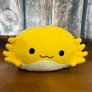 Puede incluir: Un peluche de ajolote amarillo con ojos negros de botón y una boca curvada. El juguete tiene branquias con volantes y una barriga color crema. El fondo es una superficie de madera.