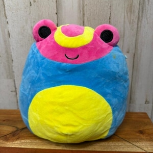 Può includere: Un giocattolo a forma di rana in peluche blu, rosa e giallo. La rana ha grandi occhi rotondi e una bocca sorridente. Il giocattolo è appoggiato su una superficie di legno.