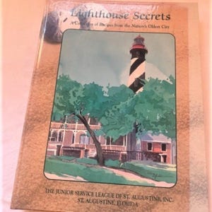 Könnte beinhalten: Ein Kochbuch mit dem Titel "Lighthouse Secrets" mit einer Aquarellillustration eines Leuchtturms und Gebäuden. Das Cover zeigt den Text "A Collection of Recipes from the Nation's Oldest City" sowie den Namen und den Ort der Organisation.