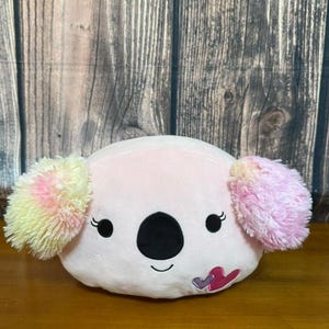 Puede incluir: Un peluche de koala rosa en forma de cojín. El koala tiene una nariz y ojos negros, con orejas esponjosas multicolores en amarillo, rosa y naranja. Un pequeño diseño en forma de corazón está en la parte inferior derecha.