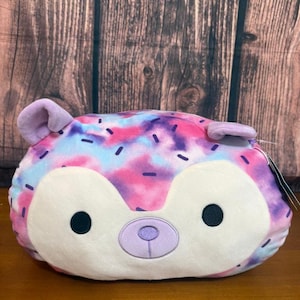 Puede incluir: Un peluche redondo con forma de erizo, con un estampado tie-dye en tonos rosa, morado y azul. El erizo tiene una cara blanca con ojos negros y una nariz morada clara. Las orejas también son de color morado claro.