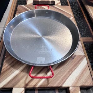 May include: A stainless steel paella pan with red handles, resting on a wooden cutting board. The pan has a circular shape with a dimpled cooking surface. The wooden board has a geometric pattern.