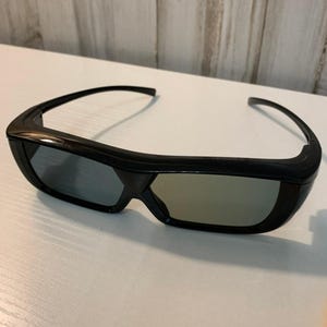 May include: Black 3D glasses featuring a thick, glossy plastic frame and rectangular lenses. The design includes curved temples that extend from the outer edges of the frame, providing a wrap-around style. The lenses have a slight greyish tint.