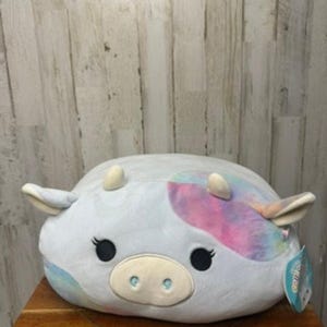 Puede incluir: Un peluche de vaca con cuerpo gris claro y parches tie-dye coloridos en rosa, azul y morado. La vaca tiene ojos negros, pestañas, una nariz rosa claro y pequeños cuernos y orejas color crema. El juguete está sobre una superficie de madera.