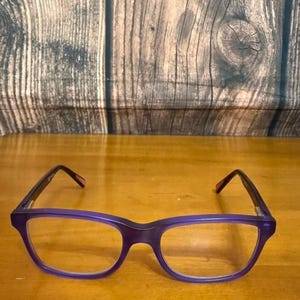 May include: A pair of purple-framed eyeglasses with clear lenses. The glasses have a rectangular shape and dark purple temples. The background is a wooden surface.