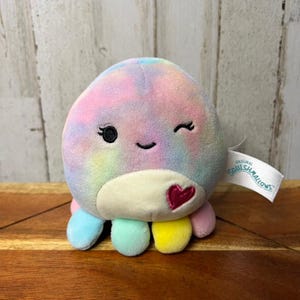 以下が含まれることがあります： パステルレインボーカラーのぬいぐるみタコのおもちゃ。おもちゃは、ウィンクする目とハート型のパッチが付いた笑顔の顔をしています。タグには「Original Squishmallows」と書かれています。