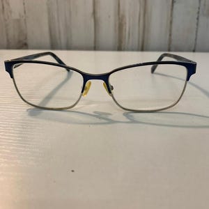 Könnte beinhalten: Eine Brille mit einem dunkelblauen oberen Rahmen und einem dünnen silbernen unteren Rand. Die Brille hat klare Gläser, einen Steg und gelbliche Nasenpads. Die Bügel sind dunkelblau und weisen ein dezentes, gesprenkeltes Muster auf.