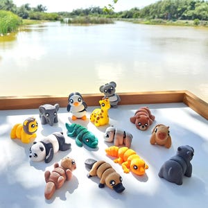 May include: A collection of colorful animal figurines, including a lion, panda, penguin, giraffe, and hippo, arranged on a white surface. The figurines are made of a soft material and are in various poses. The background features a lake and greenery.
