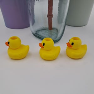 May include: Three yellow rubber ducky-shaped bottle stoppers with orange beaks and black eyes. The stoppers are in front of a clear glass bottle and two other containers.
