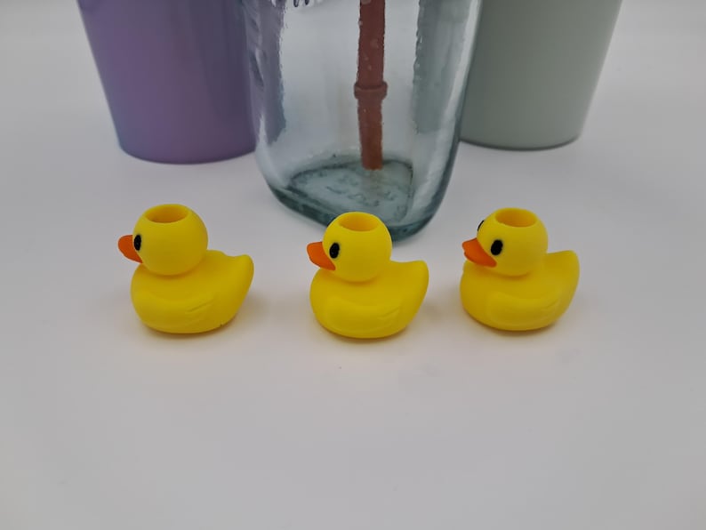 May include: Three yellow rubber ducky-shaped bottle stoppers with orange beaks and black eyes. The stoppers are in front of a clear glass bottle and two other containers.