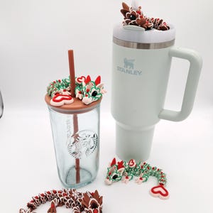 May include: A light green Stanley mug and a clear Starbucks tumbler with dragon-themed toppers and decorations. The dragons are brown, red, and green, with a gingerbread-like aesthetic. A straw is in each cup. Two additional dragon figures are also present.