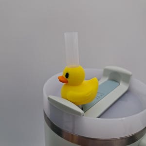 May include: A yellow rubber ducky sits atop a white and light blue Stanley tumbler lid, with a clear straw extending upwards. The duck has an orange beak and black eyes. The tumbler is light green with a silver rim.