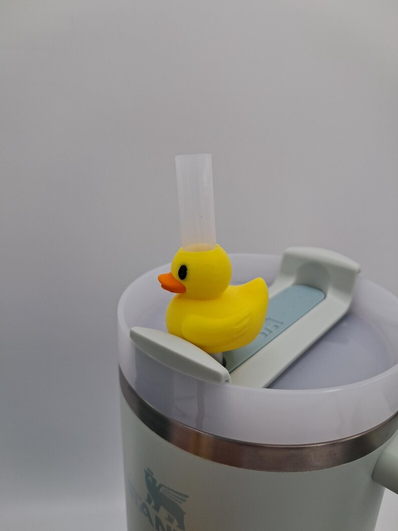 May include: A yellow rubber ducky sits atop a white and light blue Stanley tumbler lid, with a clear straw extending upwards. The duck has an orange beak and black eyes. The tumbler is light green with a silver rim.
