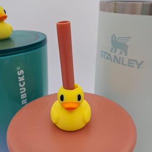 May include: A yellow duck-shaped straw topper with a brown straw inserted into its head. The duck has black eyes and an orange beak. The topper sits on a brown lid, with a teal and white tumbler and a white Stanley tumbler in the background.