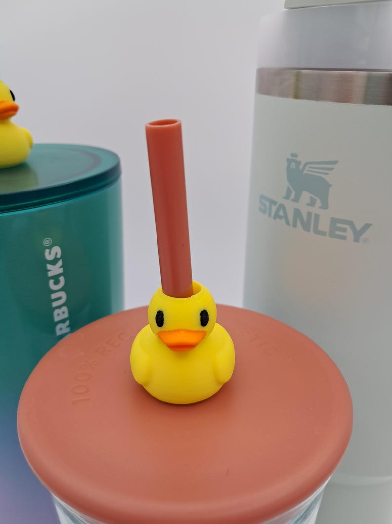 May include: A yellow duck-shaped straw topper with a brown straw inserted into its head. The duck has black eyes and an orange beak. The topper sits on a brown lid, with a teal and white tumbler and a white Stanley tumbler in the background.