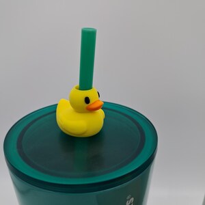 May include: A teal tumbler with a yellow duck-shaped straw topper and a green straw. The duck has an orange beak and black eyes. The tumbler has the Starbucks logo on the side.
