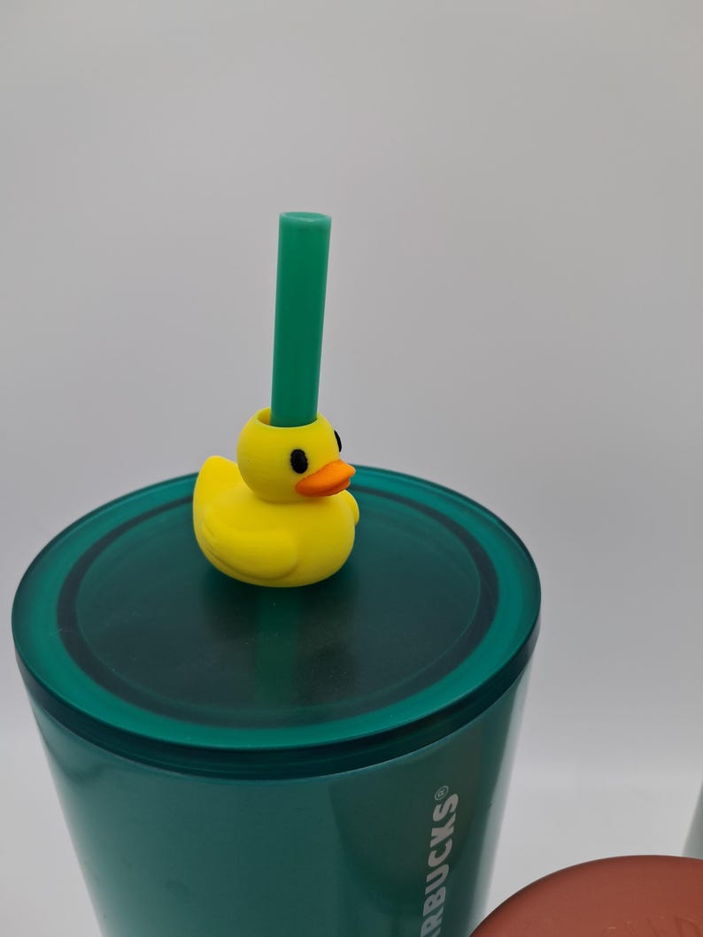 May include: A teal tumbler with a yellow duck-shaped straw topper and a green straw. The duck has an orange beak and black eyes. The tumbler has the Starbucks logo on the side.