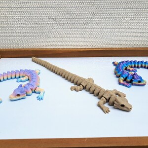 May include: Three articulated lizard figures in a wooden tray. One is tan, the others are rainbow-colored. The lizards are posed on a white surface, with a textured gray background.