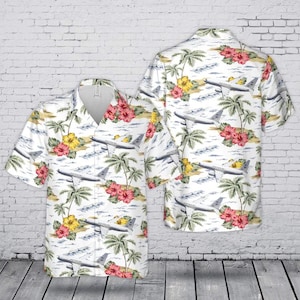 May include: A short-sleeve button-down shirt featuring a white background with an all-over print of airplanes, green palm trees, and red and yellow hibiscus flowers. The shirt has a classic collar and a front button closure with a straight hem.