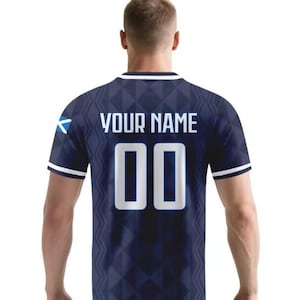 May include: A dark blue short-sleeved jersey featuring a geometric diamond pattern, white trim on the collar and sleeves, and a small Scottish flag on the left sleeve. The back displays the text YOUR NAME above the large white numbers 00.