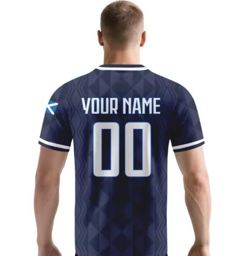 May include: A dark blue short-sleeved jersey featuring a geometric diamond pattern, white trim on the collar and sleeves, and a small Scottish flag on the left sleeve. The back displays the text YOUR NAME above the large white numbers 00.