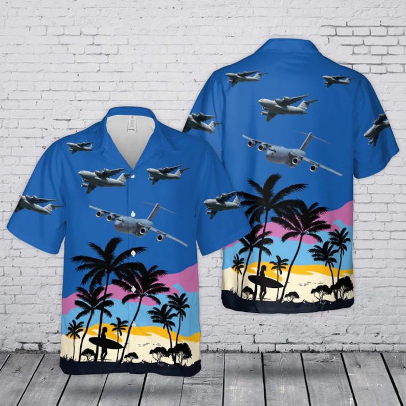 Puede incluir: Una camisa de manga corta con botones en color azul que presenta un patr&oacute;n repetitivo de aviones de transporte militar sobre un paisaje tropical con siluetas negras de palmeras, un cielo al atardecer y una persona sosteniendo una tabla de surf.