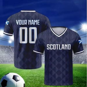 May include: A dark blue football shirt with a geometric diamond pattern, white trim on the V-neck and sleeves, and a small Scottish flag on the sleeve. The front displays the text SCOTLAND, while the back features the text YOUR NAME and 00.
