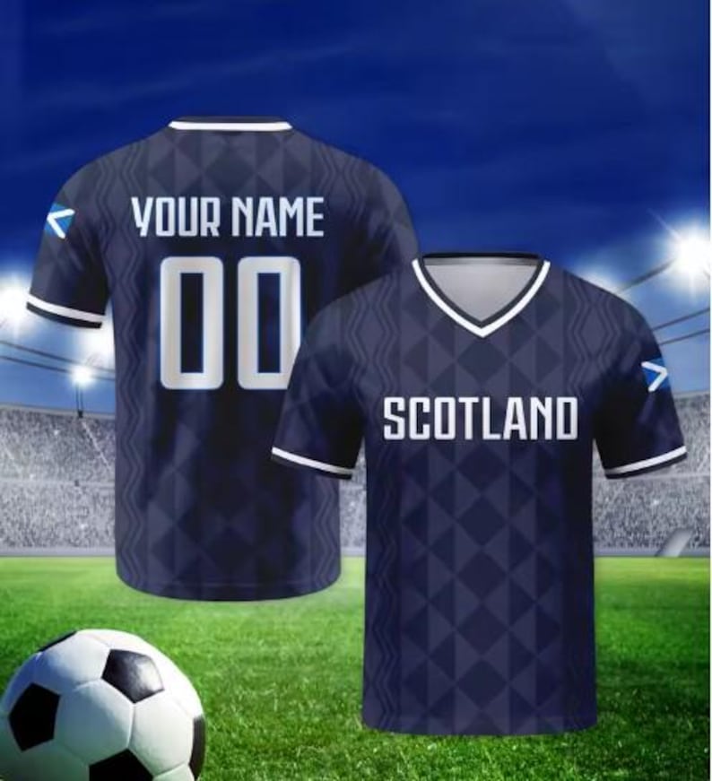 May include: A dark blue football shirt with a geometric diamond pattern, white trim on the V-neck and sleeves, and a small Scottish flag on the sleeve. The front displays the text SCOTLAND, while the back features the text YOUR NAME and 00.
