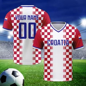 May include: Two Croatia football shirts with red and white chequered patterns, the text "YOUR NAME" and the number "00" on the back. The other shirt has "CROATIA" printed on the front. A football rests on the green grass.
