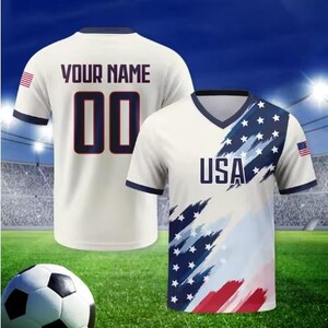 May include: A white V-neck jersey featuring a blue and red brushstroke pattern with white stars, the text USA, and a small American flag on the sleeve. The back displays the text YOUR NAME and the number 00 in dark blue with a red outline.