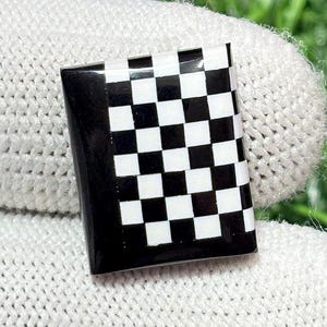 Może przedstawiać: Mały, prostokątny przedmiot z czarno-białym wzorem w szachownicę. Wzór przedstawia powtarzający się wzór czarnych i białych kwadratów. Przedmiot ma błyszczące wykończenie i prawdopodobnie jest akcesorium dekoracyjnym.