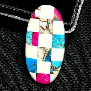 Pode incluir: Uma pedra preciosa oval com um padrão xadrez. O padrão alterna entre branco, turquesa, rosa e um marmoreado castanho e dourado. A pedra preciosa está sobre um fundo preto.