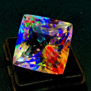 Könnte beinhalten: Ein großer, quadratisch geschliffener Edelstein mit einer schillernden Farbpalette, darunter Blau-, Grün-, Rot- und Gelbtöne. Der Edelstein ist vor einem schwarzen Hintergrund platziert, der seine facettenreiche Oberfläche und die brillante Lichtbrechung hervorhebt. Schmuck.