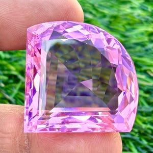 Könnte beinhalten: Ein facettierter, halbkreisförmiger Edelstein in Rosa. Der Stein wird zwischen Fingern gehalten, was seinen komplizierten Schliff und seine reflektierenden Oberflächen hervorhebt. Der Hintergrund ist ein verschwommenes Grün, das mit der Farbe und den Details des Steins kontrastiert.