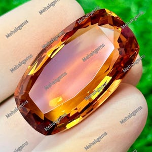 Puede incluir: Una piedra preciosa de corte ovalado con un degradado vibrante en naranja y amarillo dorado, que presenta múltiples facetas que reflejan la luz. La piedra muestra una superficie pulida y translúcida con el texto Mehakgems repetido.