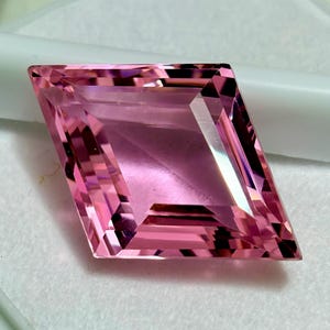Puede incluir: Una gema rosa facetada con forma de cometa. La gema tiene múltiples facetas que reflejan la luz, creando un efecto brillante. El color es un rosa suave y rosado, y la gema parece estar sobre un fondo blanco.
