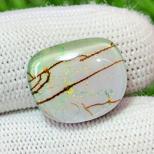 May include: A polished, oval gemstone with a translucent, pale green top and a white base. The stone has intricate brown vein-like patterns and iridescent flashes of colour, creating a unique and natural appearance.