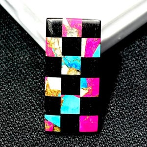 Puede incluir: Una piedra preciosa rectangular con un patrón de tablero de ajedrez en cuadrados negros, blancos, dorados, turquesas y rosas. La piedra preciosa está sobre un fondo negro texturizado y una superficie blanca.