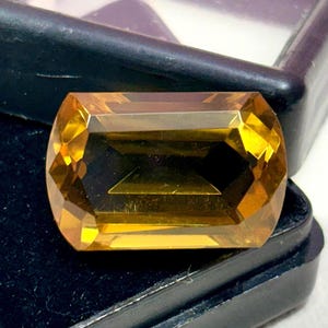 以下が含まれることがあります： 温かみのある黄金色の長方形カットの宝石。黒い背景に配置され、その透明度と輝きを際立たせています。カットは複数のファセットを備え、光を反射してきらめく効果を生み出しています。