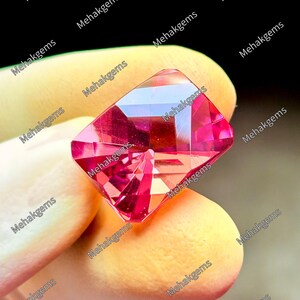 Puede incluir: Una piedra preciosa rectangular facetada con un tono rosa y rojizo vibrante. La piedra presenta un corte geométrico complejo con múltiples facetas triangulares y trapezoidales que reflejan la luz. El texto Mehakgems está superpuesto repetidamente en la imagen.