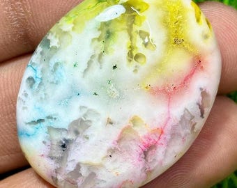 May include: An oval gemstone displaying a blend of colours. The stone has a white base with streaks of yellow, green, blue, and pink. The colours merge to create a unique, marbled effect. The gemstone is held in a hand.