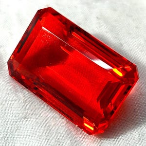 Può includere: Una gemma rossa rettangolare sfaccettata, con bordi affilati e una superficie lucida. La gemma è posta su uno sfondo bianco strutturato. Il colore della gemma è un rosso vibrante e trasparente.