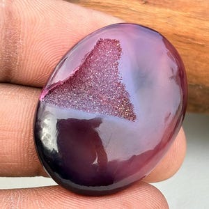 Può includere: Una gemma ovale lucidata, di colore viola intenso. La pietra presenta una cavità riempita di formazioni cristalline scintillanti, rosa e oro. La superficie riflette la luce, creando riflessi.