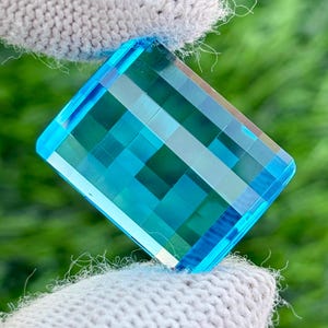 Peut inclure: Une pierre précieuse rectangulaire et facettée d'un bleu vif. La surface est divisée en une grille de petites facettes carrées, créant un jeu d'ombre et de lumière. Le bijou est tenu par un gant en tricot gris clair, avec un arrière-plan vert flou.