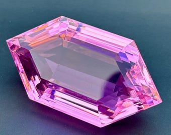 May include: A faceted, rectangular gemstone with a vibrant pink hue. The gem's cut reveals multiple facets, creating a play of light and shadow. The gemstone is set against a dark background, highlighting its color and clarity.