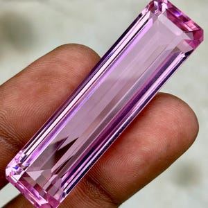 May include: A rectangular, faceted gemstone in shades of pink and lavender. The gem has a long, slender shape with precise cuts and polished surfaces, reflecting light. The gemstone is held between fingers, showcasing its color and clarity.