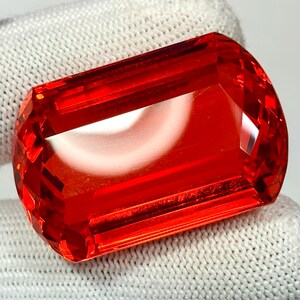 May include: A faceted, rectangular-cut gemstone with a vibrant, fiery red hue. The gem is held against a white, textured fabric, showcasing its clarity and brilliance. The cut of the stone highlights its internal structure and light refraction.